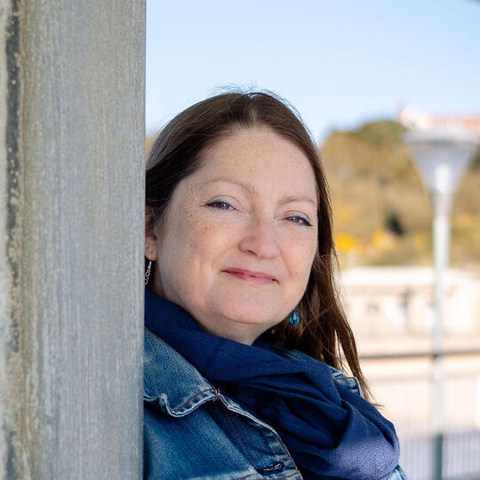 Bronwen Leigh is smiling to camera, leans against a wooden post wearing a blue denim jacket and scarf.